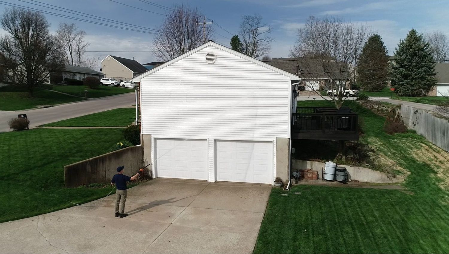 House Exterior Inspection A man soft washing a white, two-car garage attached to a house. The home sits on a green lawn with a deck visible on the right side. The scene highlights a benefit of soft washing for home exterior cleaning.