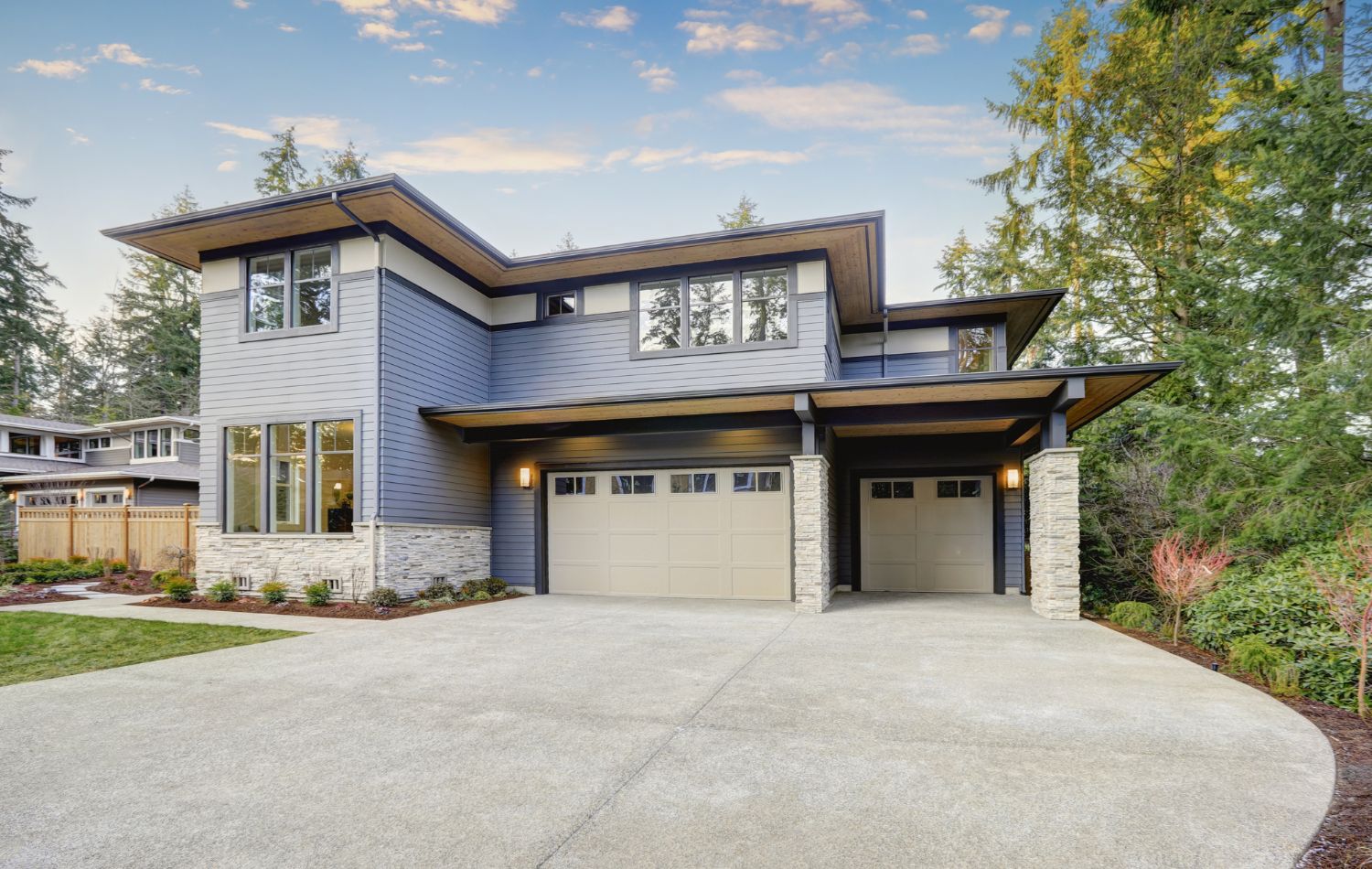 Modern two-story home with light blue siding, stone accents, and a two-car garage. A concrete driveway leads up to the house, which is surrounded by lush greenery and trees.
