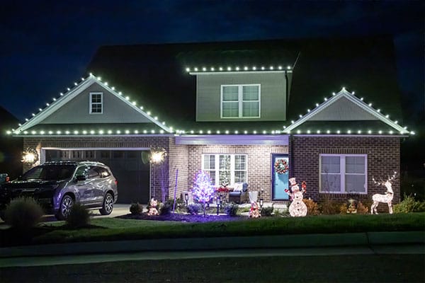 Christmas Light Service House decorated with Christmas lights on the roofline and festive decorations in the yard, including a snowman, reindeer, and illuminated Christmas tree. A Christmas light service ensures a beautiful holiday display.
