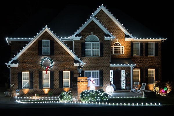 Brick home illuminated with bright white Christmas lights outlining the roof, a festive wreath on the front, and cheerful holiday decorations in the yard. Hiring a Christmas light company ensures a stunning display.