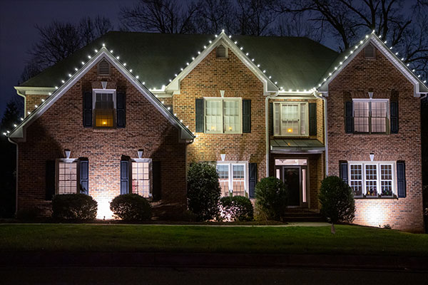 Two-story brick house at night, professionally decorated with bright white holiday lights along the roofline. Dark shutters accent the windows. Green bushes line the manicured lawn.