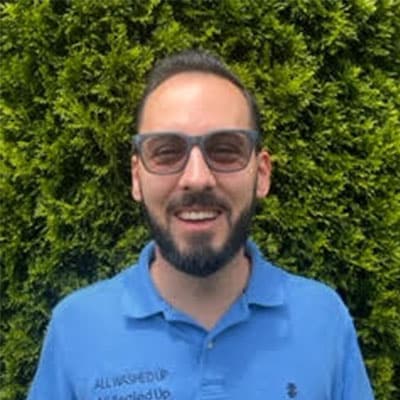 Man with a beard and sunglasses smiles in front of a green hedge. He's wearing a blue polo shirt with "All Washed Up" printed on it.