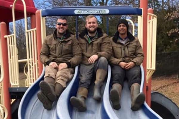 Three men wearing matching brown jackets and boots sit side-by-side on a blue and white playground slide on a sunny day.