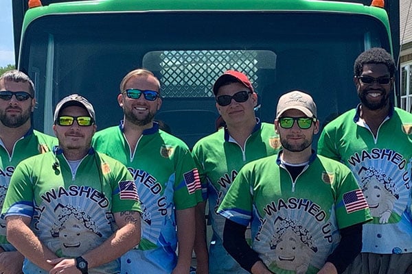 Group of six men wearing matching green shirts with "All Washed Up" text and a sheep graphic, standing in front of a green truck. American flags are visible on the sleeves of their shirts.