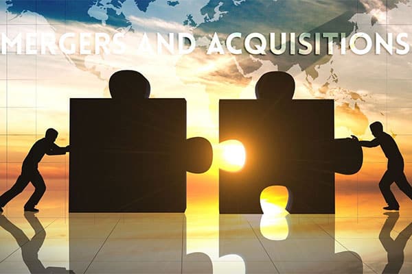 Silhouetted figures push large puzzle pieces together against a sunset backdrop, symbolizing mergers and acquisitions. The pieces connect, revealing bright light, with a world map above.