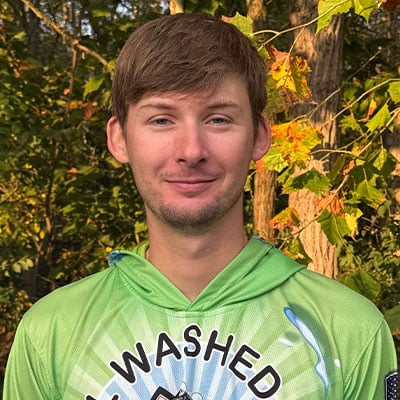 Portrait of a young man in a green "Washed" hoodie, smiling slightly. He is outdoors with trees and foliage in the background.
