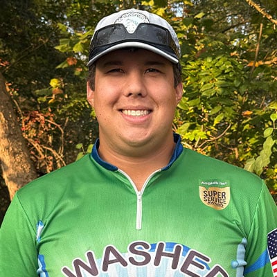 Smiling man in a green fishing shirt with "Washed" printed on it, wearing a cap with sunglasses perched on top. He stands outdoors with trees in the background.