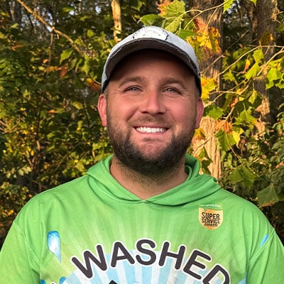 Smiling man in a green "Washed" hoodie and baseball cap stands outdoors. The hoodie has a "Super Service Award" badge. Autumn leaves provide a colorful backdrop.