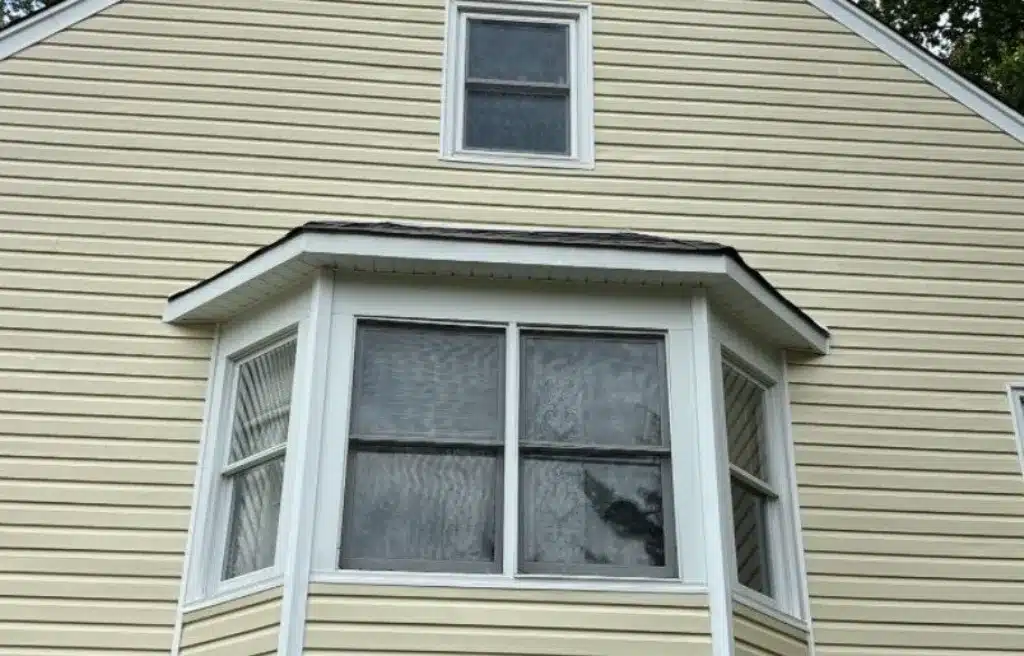 Exterior view of a home with light yellow vinyl siding and white-framed bay windows, showcasing the need for removing winter residue. A small square window is visible above the bay window.