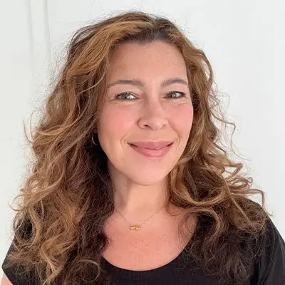 Smiling woman with long wavy light brown hair, wearing a black top and small gold necklace, against a plain white background.