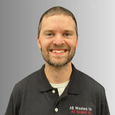 Smiling person with short hair and beard wearing black polo embroidered All Washed Up All Sealed Up on gray gradient background.